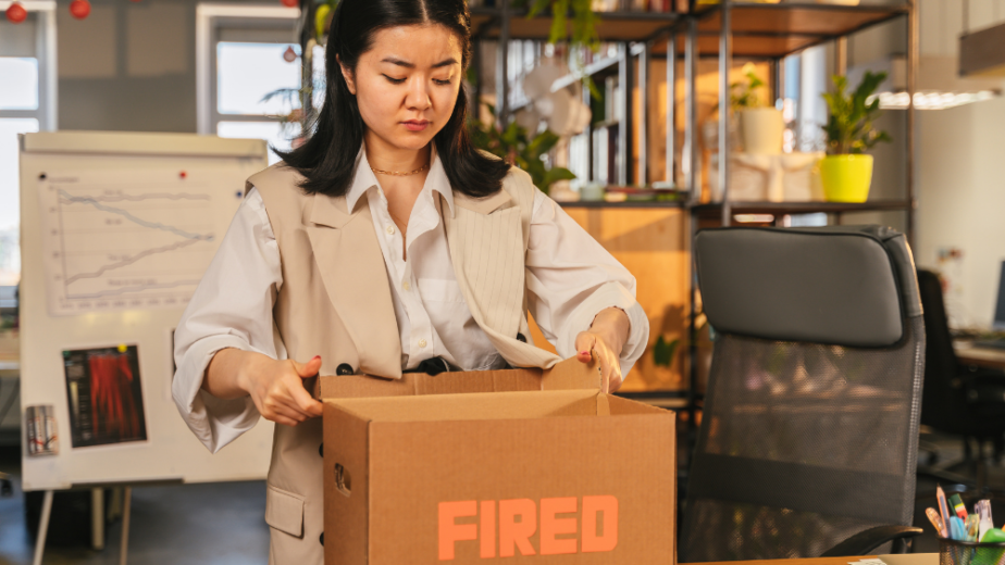 a female asian woman opening a brown box with orange words on the front spelled out "FIRED" in her office