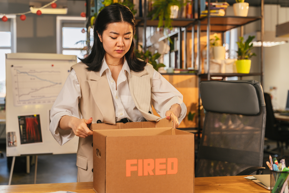 a female asian woman opening a brown box with orange words on the front spelled out "FIRED" in her office