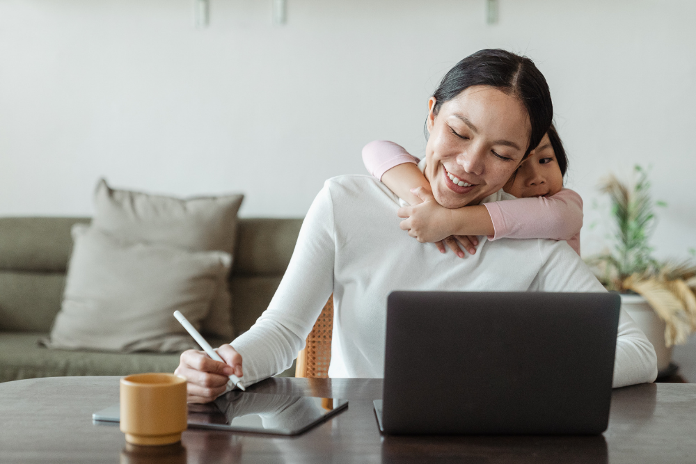 a mom with her child hugging her from behind while she is working on the laptop
