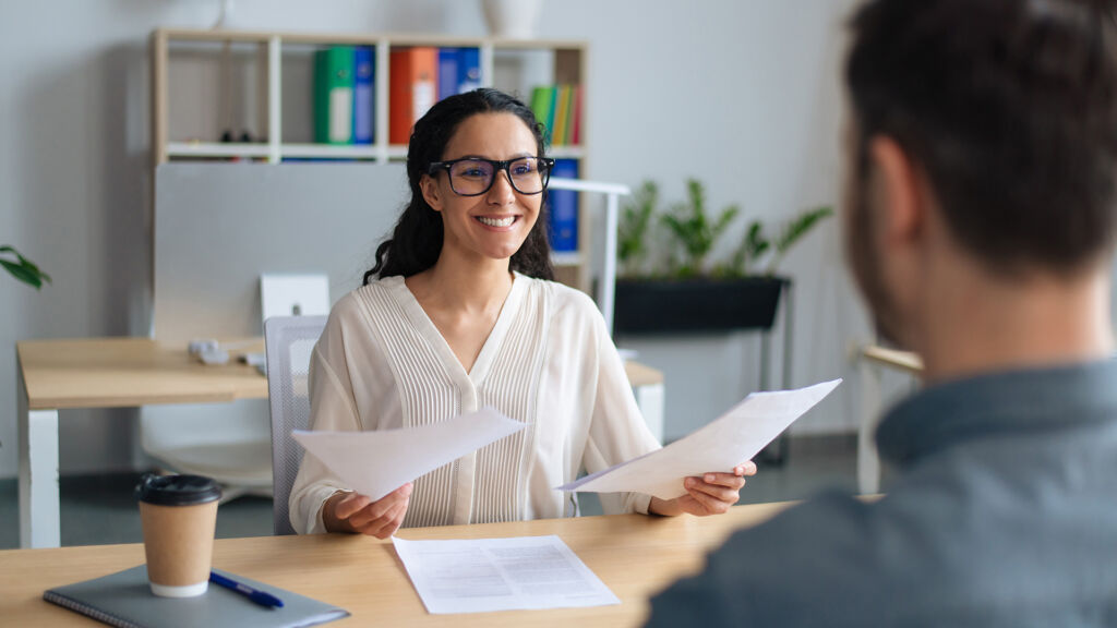 a smiling woman with glasses holding papers in both hands and looking at a man sitting in front of her